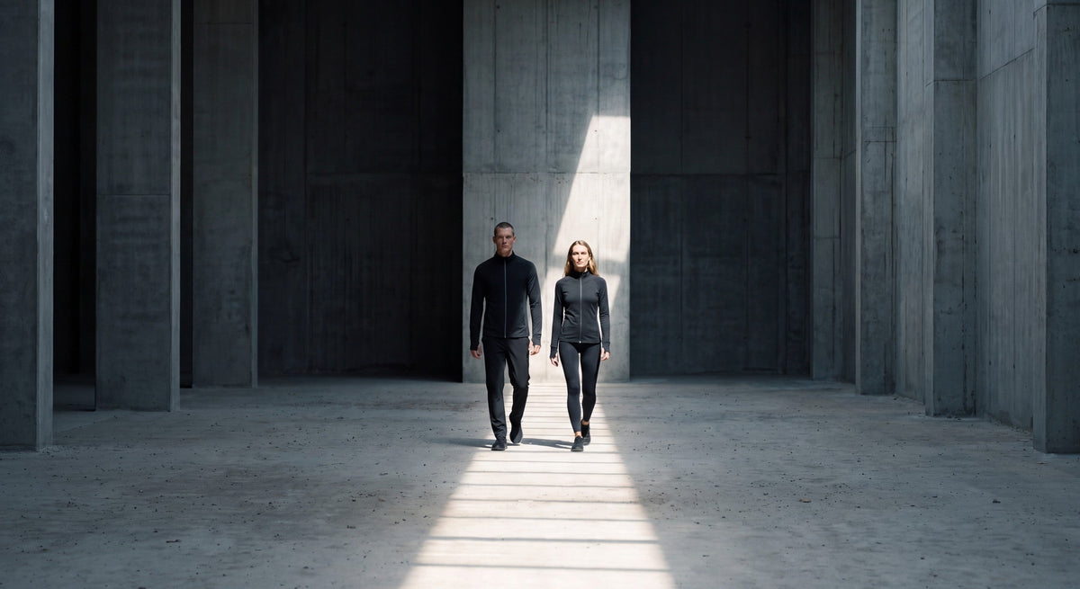 Two people in black track jackets walking through a sunlit concrete hall